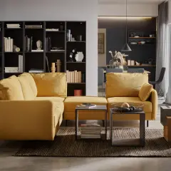 Sofa w odcieniu **kolor musztardowy** przyciąga wzrok. Obok niej regał z książkami i stoliki kawowe.