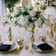 Eleganckie nakrycie stołu z białymi kwiatami, złotymi buteleczkami i kieliszkami.