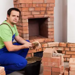 Murarz w niebieskich ogrodniczkach i zielonej koszulce układa cegły szamotowe, budując kominek.