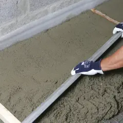 Ręce w rękawiczkach wyrównują świeżą wylewkę budowlaną długą łopatką.