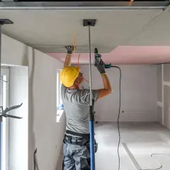 Budowlaniec w żółtym kasku montuje płyty nida gips na suficie, używając wiertarki i podpór.