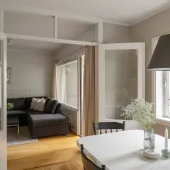 Jasny salon z szarą kanapą i stołem, na którym stoją świece i wazon z kwiatami. W tle widać okna i drzwi, a na ścianach subtelne wzory geometryczne.