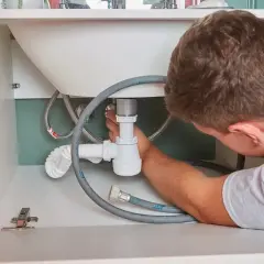 Instalator pracuje pod umywalką, podłączając syfon. Sprawdź montaż umywalki cena, by poznać koszty instalacji.