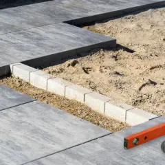 Budowa tarasu betonowego w toku. Betonowe płyty i obrzeża czekają na ułożenie. Poziomica i młotek gumowy gotowe do pracy.