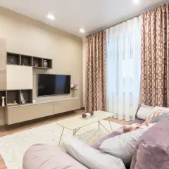Nowoczesny salon z sofą w kolorze fioletowym. Ściany w odcieniach **kolor beżowy a kremowy**, meble RTV i stolik kawowy również w tych barwach. Zasłony z geometrycznym wzorem.