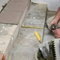 Płytkarz mierzy taśmą podłogę przed układaniem płytek. Widać też kielnie i wiadro z zaprawą.