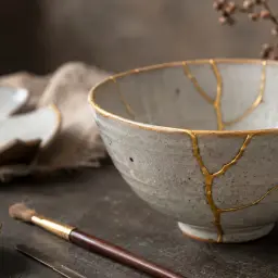 Ceramiczna miska i talerze z widocznymi złotymi pęknięciami, świadectwo sztuki kintsugi. Obok pędzel i złoty pył.