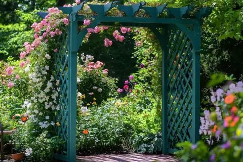 Zielona pergola DIY, ozdobiona kwitnącymi różami i innymi kwiatami, tworzy malownicze przejście do ogrodu pełnego zieleni.