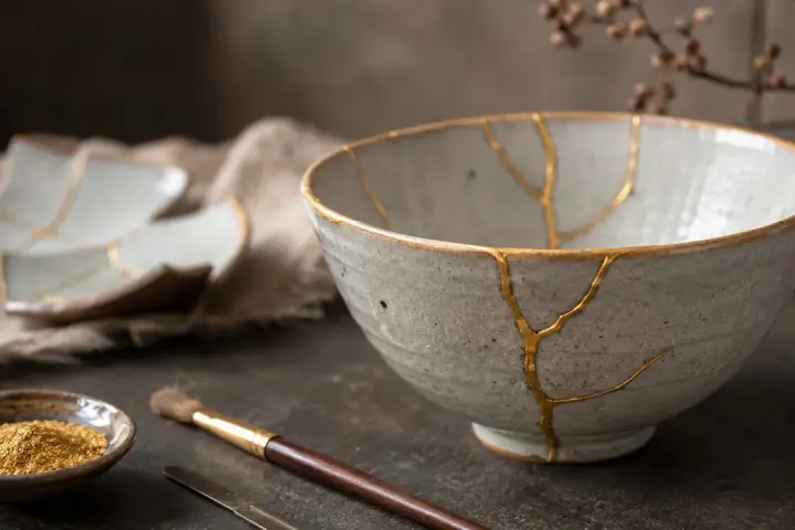 Ceramiczna miska i talerze z widocznymi złotymi pęknięciami, świadectwo sztuki kintsugi. Obok pędzel i złoty pył.
