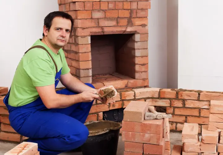 Murarz w niebieskich ogrodniczkach i zielonej koszulce układa cegły szamotowe, budując kominek.