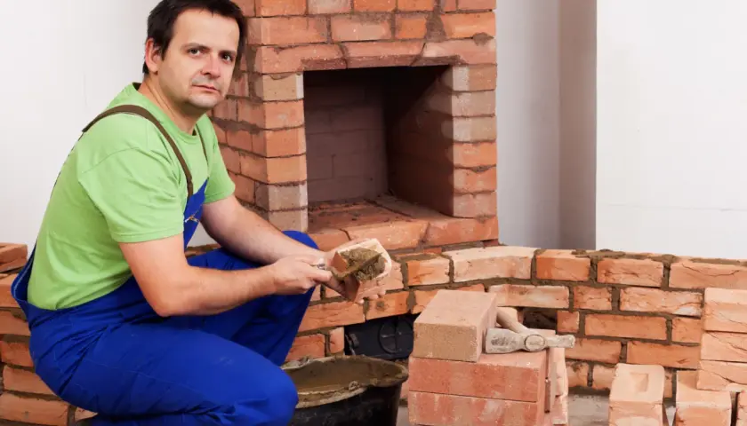 Murarz w niebieskich ogrodniczkach i zielonej koszulce układa cegły szamotowe, budując kominek.