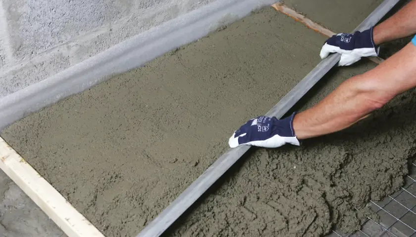 Ręce w rękawiczkach wyrównują świeżą wylewkę budowlaną długą łopatką.