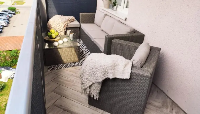 Przytulny balkon w bloku z wielkiej płyty, z meblami rattanowymi, kocami i stolikiem kawowym.