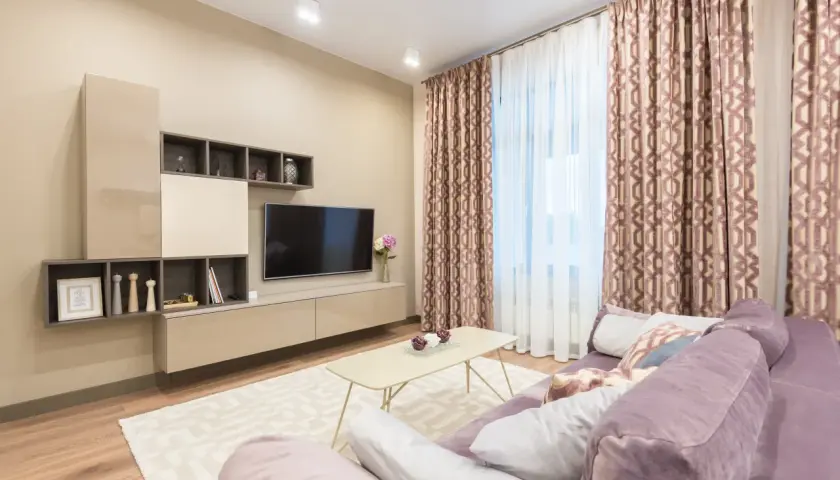 Nowoczesny salon z sofą w kolorze fioletowym. Ściany w odcieniach **kolor beżowy a kremowy**, meble RTV i stolik kawowy również w tych barwach. Zasłony z geometrycznym wzorem.
