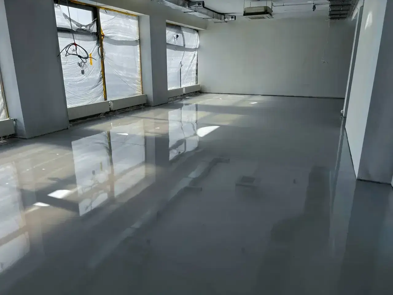 Jasne, lśniące podłogi epoksydowe odbijają światło w pustym pomieszczeniu z oknami zakrytymi folią.