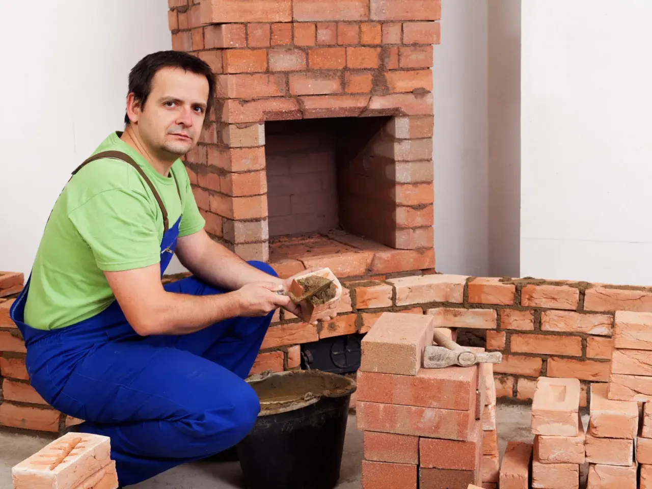 Murarz w niebieskich ogrodniczkach i zielonej koszulce układa cegły szamotowe, budując kominek.