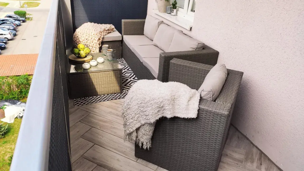Przytulny balkon w bloku z wielkiej płyty, z meblami rattanowymi, kocami i stolikiem kawowym.