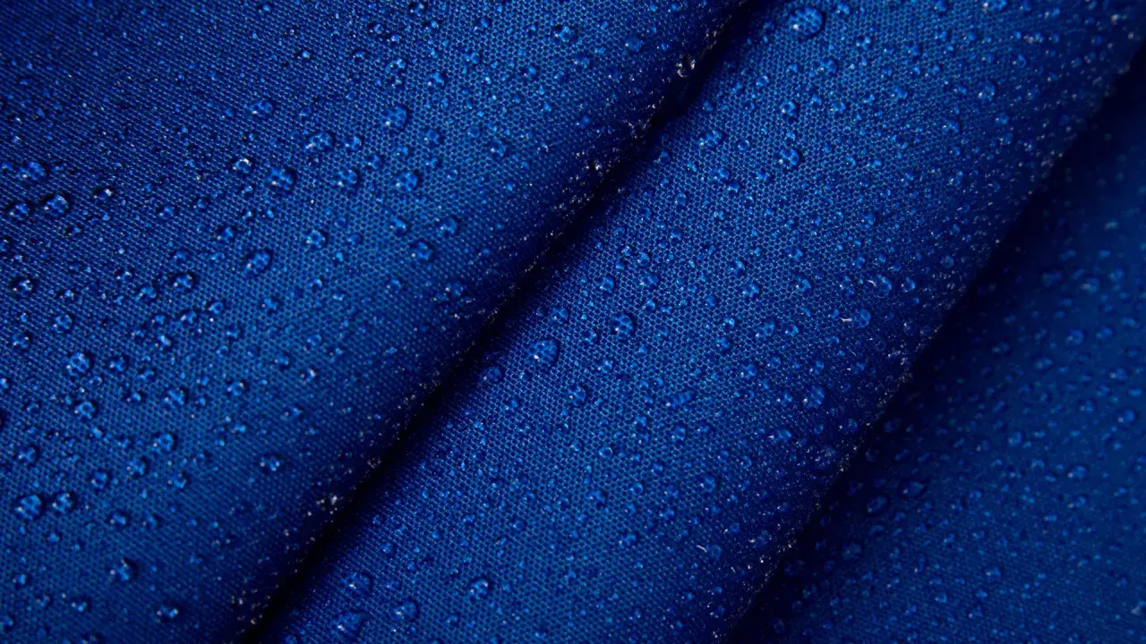 Krople wody na granatowym, **hydrofobowym** materiale. Tekstura tkaniny jest wyraźnie widoczna pod kropelkami.
