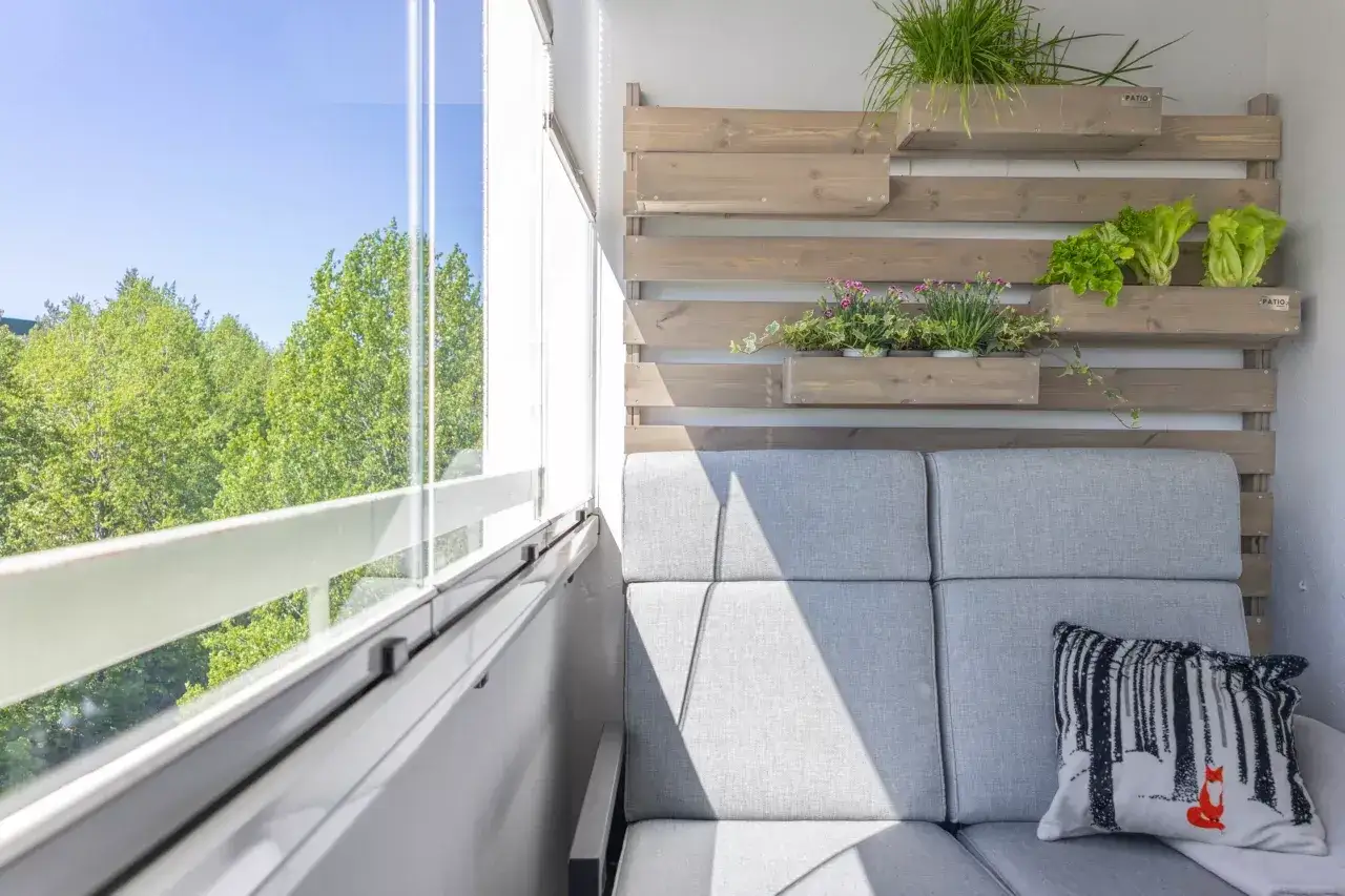 Przytulny balkon w bloku z wielkiej płyty, z szarą sofą, zielenią i widokiem na drzewa.