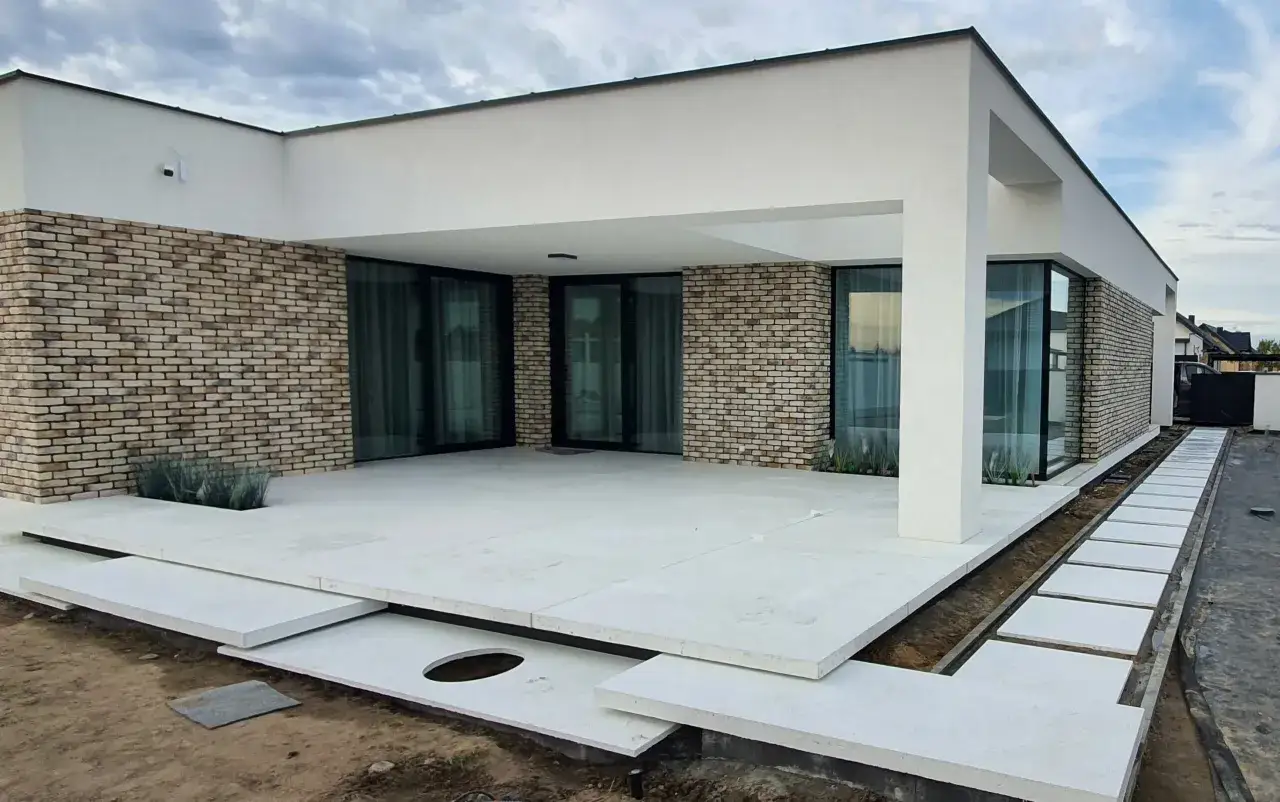 Nowoczesny dom z ceglaną ścianą i przestronnym tarasem betonowym. Ścieżka z płyt prowadzi do wejścia.